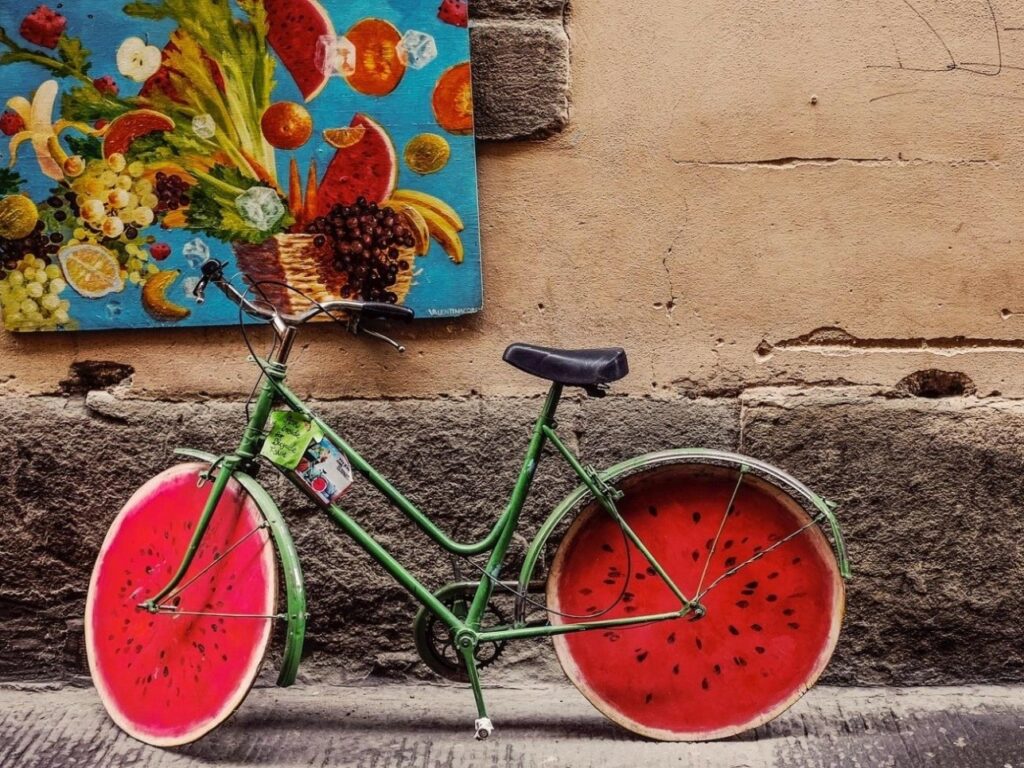 Grünes Fahrrad lehnt an einer Hauswand, mit Rädern wie Wassermelonenscheiben; daneben buntes Obstbild an der Wand.