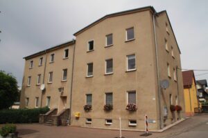 Ein beiges dreistöckiges Wohnhaus mit vielen Fenstern, Satellitenschüsseln und Blumenkästen an den Fenstern.