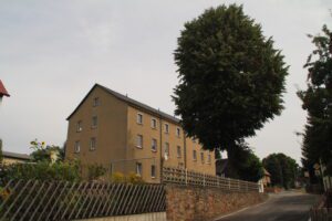 Mehrstöckiges beiges Wohnhaus an einer Straße, mit Steinmauer, Gartenzaun und großem Baum im Vordergrund.