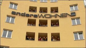 Mehrere Menschen winken aus den geöffneten Loggien eines gelben Wohnhauses mit dem Schriftzug ‚andersWohnen eG‘.