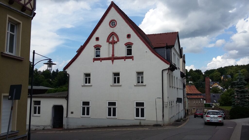 Weißes historisches Haus mit rotem Giebeldekor an einer kurvigen Straße in einer kleinen Stadt.