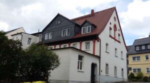 Großes weißes Wohnhaus mit rotem Dach und dunklem Schiefergiebel, teils verdeckt von Büschen.