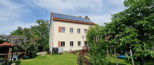 Ein Wohnaus mit Solarpanelen auf dem Dach, umgeben von grünem Garten, Bäumen und blauem Himmel.