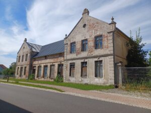 Verfallenes zweigeteiltes Gebäude mit bröckelnder Fassade, beschädigten Fenstern und verwachsenem Umfeld an einer Straße.