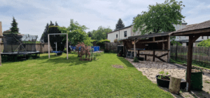 Großer Garten mit Trampolin, Schaukel, Pool und überdachtem Sitzbereich, umgeben von Bäumen und Rasen.