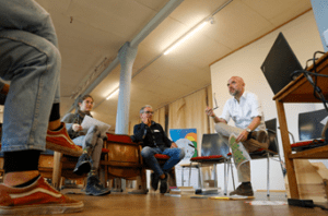 Mehrere Personen sitzen in einem hellem Raum im Kreis zusammen. Eine Person spricht.