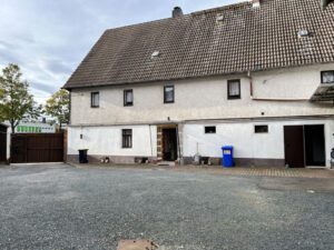 Großer Innenhof mit Schotterfläche vor einem langem Wohngebäude mit Satteldach, Fenstern und mehreren Türen.