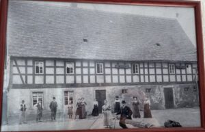 Altes Schwarzweißfoto eines langen Fachwerkhauses, davor eine Gruppe von Menschen in historischer Kleidung.