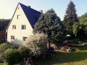 Ein helles Wohnhaus mit großem Garten, Bäumen, Hängematte, Fahrrädern und Menschen an einem Gartentisch.
