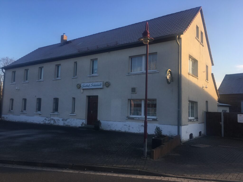 Alter Gasthof mit heller Fassade, roten Dachziegeln und Schild ‚Gasthof Schützehof‘ im Abendlicht.