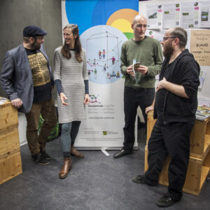 Vier Personen stehen vor einem Banner der Dezentrale – Netzwerk für gemeinschaftliches Wohnen.