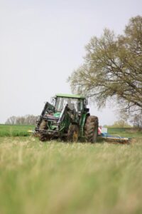 Grüner Traktor arbeitet auf einem Feld. Am Rand steht ein großer Baum. Der Himmel ist bewölkt.