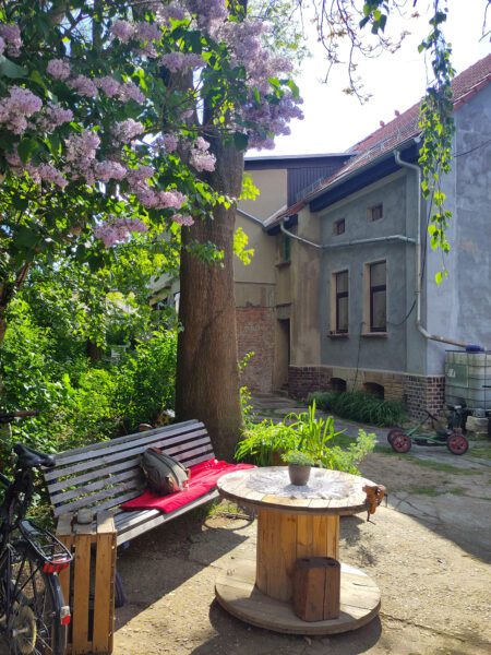 Gemütliche Sitzecke im Hof mit Holzbank, Tisch aus Kabeltrommel unter einem blühendem Fliederbaum.