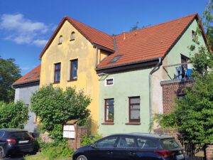 Buntes Wohnhaus mit rotem Dach, blauer, gelber und grüner Fassade, Autos parken davor.