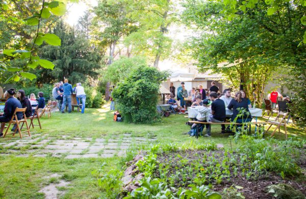 Menschen sitzen und stehen an Tischen im sonnigen Garten, essen und unterhalten sich.