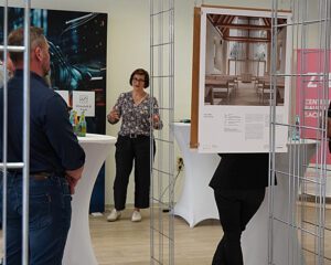 Mehrere Personen stehen in einer Ausstellung, eine Frau spricht vor aufgehängten Architekturfotos.