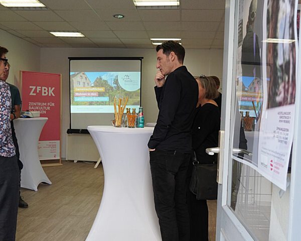 Menschen stehen an Stehtischen in einem Ausstellungsraum, im Hintergrund läuft eine Präsentation.