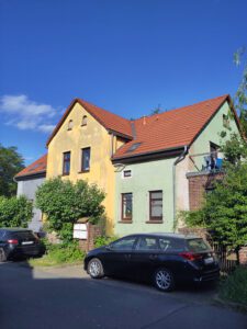 Buntes Wohnhaus mit rotem Dach, blauer, gelber und grüner Fassade, Autos parken davor.