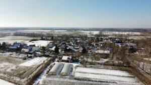 Winterliche Luftaufnahme eines Dorfes mit schneebedeckten Feldern, Gewächshäusern und verstreuten Häusern.