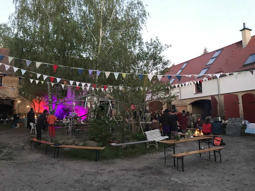 Abendliche Hofszene mit bunten Wimpeln, Lichtern und Menschen, die gemeinsam feiern und zusammen sitzen.