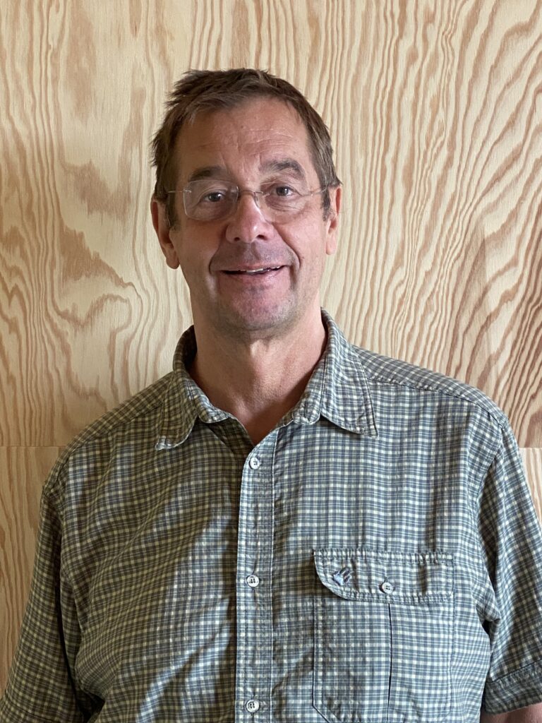 Ein Porträtfoto von Matthias Voigt. Er schaut in die Kamera und steht vor einem Hintergrund in Holzoptik.