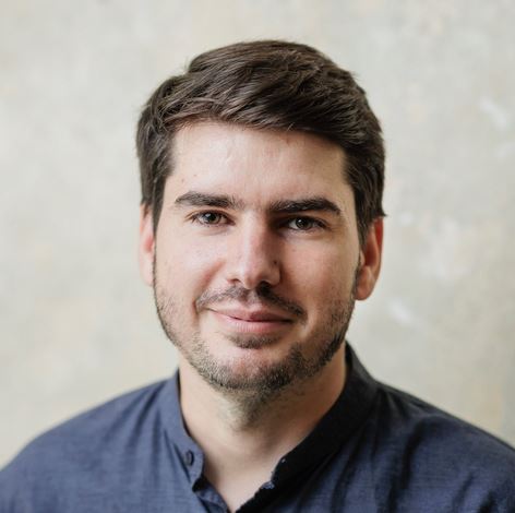 Ein Porträtfoto von Sandro Kühn. Er schaut in die Kamera. Der Hintergrund ist hell.