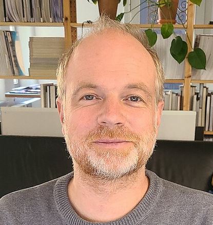 Ein Porträtfoto von Winfried Endres. Er schaut in die Kamera. Im Hintergrund ist ein Regal mit Büchern.