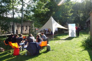 Eine Gruppe Menschen sitzt bei sonnigem Wetter in einem Stuhlkreis auf einer Wiese und unter einem Tarp und tauschen sich aus.