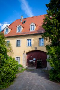 Ein gelbes Gebäude mit rotem Tor und Dach, umgeben von Bäumen und Sträuchern unter blauem Himmel.