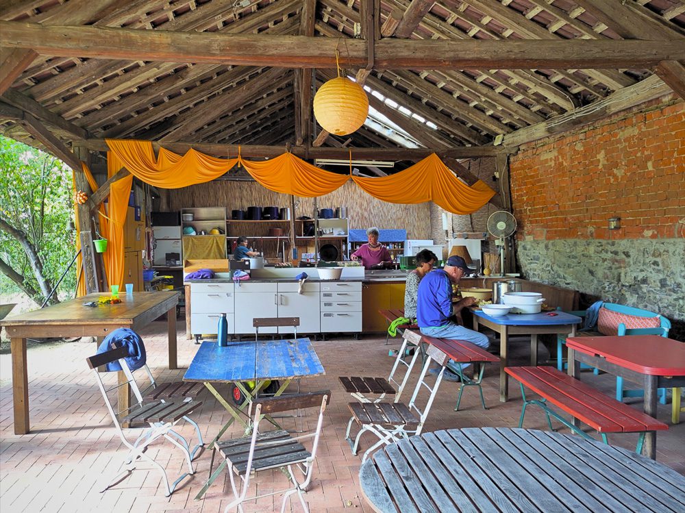 Offene Sommerküche mit Holzdach, gelben Stoffbahnen, Tischen und Menschen beim gemeinsamen Kochen.