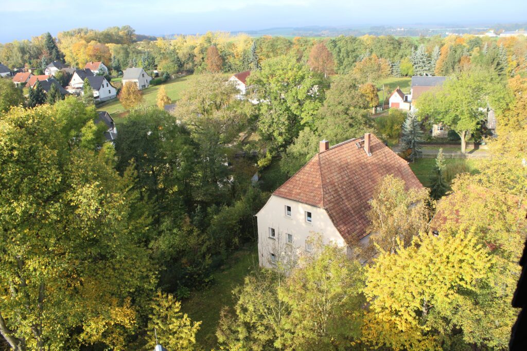 Blick von oben auf das Pfarrhaus in einer herbstlich grünen Landschaft. Im Vordergrund steht das helle Wohnhaus mit rotem Ziegeldach, umgeben von dichtem, teils herbstlich verfärbtem Laub. Im Hintergrund liegen verstreut weitere Häuser mit Gärten, Felder und ein breiter Streifen Wald. Am Horizont ist die weite Landschaft unter einem leicht bewölkten Himmel zu erkennen.
