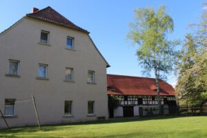 Ein freundliches Bauernhofensemble im Sonnenschein. Im Vordergrund eine gepflegte grüne Wiese. Links ein zweistöckiges, beiges Wohnhaus mit mehreren Fenstern, rechts davon ein Fachwerkgebäude mit rotem Ziegeldach. Zwischen den Gebäuden steht eine schlanke Birke mit frischem Laub. Der Himmel ist klar blau.