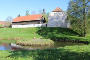 Zwei Gebäude stehen auf einer grünen Anhöhe an einem kleinen Teich. Links ein langes Gebäude mit dunklem Holz und rotem Ziegeldach, rechts ein helles, mehrstöckiges Wohnhaus. Im Vordergrund wächst grünes Gras mit Löwenzahnblüten. Dazwischen ist ein Teich mit einer kleinen Insel zu sehen. Im Hintergrund sind Bäume und der Turm einer Kirche mit Zwiebeldach zu sehen, der Himmel ist klar blau.