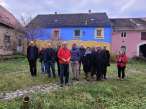 Eine Gruppe von Menschen steht auf einer Wiese in einem Innenhof vor bunt bemalten Gebäuden in Blau, Gelb und Rosa.