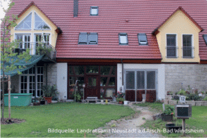 Modernisiertes Wohnhaus mit rotem Satteldach, großen Fenstern und Wintergarten. Vor dem Haus liegt ein Garten mit Rasen, Pflanzen und einem Weg zum Eingang.